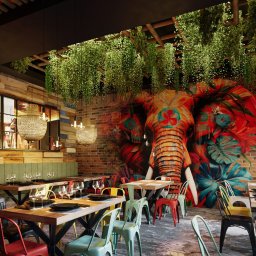 REDLICKI STUDIO Piotr Redlisz-Redlicki - Restauracja z muralem słonia na ceglanej ścianie, wiszącymi roślinami i kolorowymi krzesłami. Widoczne nakryte stoły i lampy w stylu industrialnym.