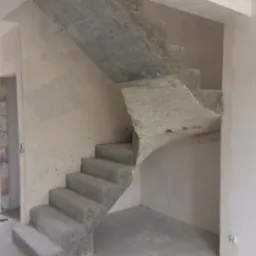 Surowe, betonowe schody spiralne w jasnym wnętrzu w stanie deweloperskim. Widoczne ślady szalunku i niedoskonałości betonu, podkreślające industrialny charakter.