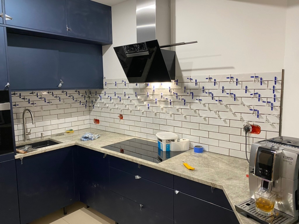 Układanie białych, prostokątnych płytek na ścianie w kuchni. Widoczne niebieskie krzyżyki dystansowe, ciemnoniebieskie szafki i ekspres do kawy.