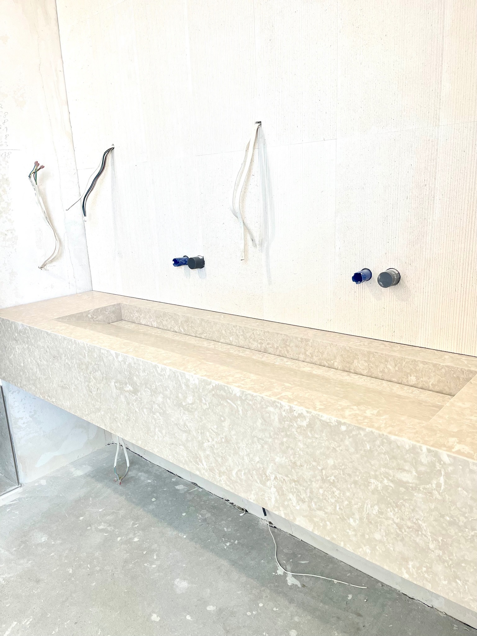 Beżowa, marmurkowa umywalka wpuszczana w blat, zamontowana na ścianie z widocznymi przewodami i instalacją hydrauliczną w surowym wnętrzu łazienki.