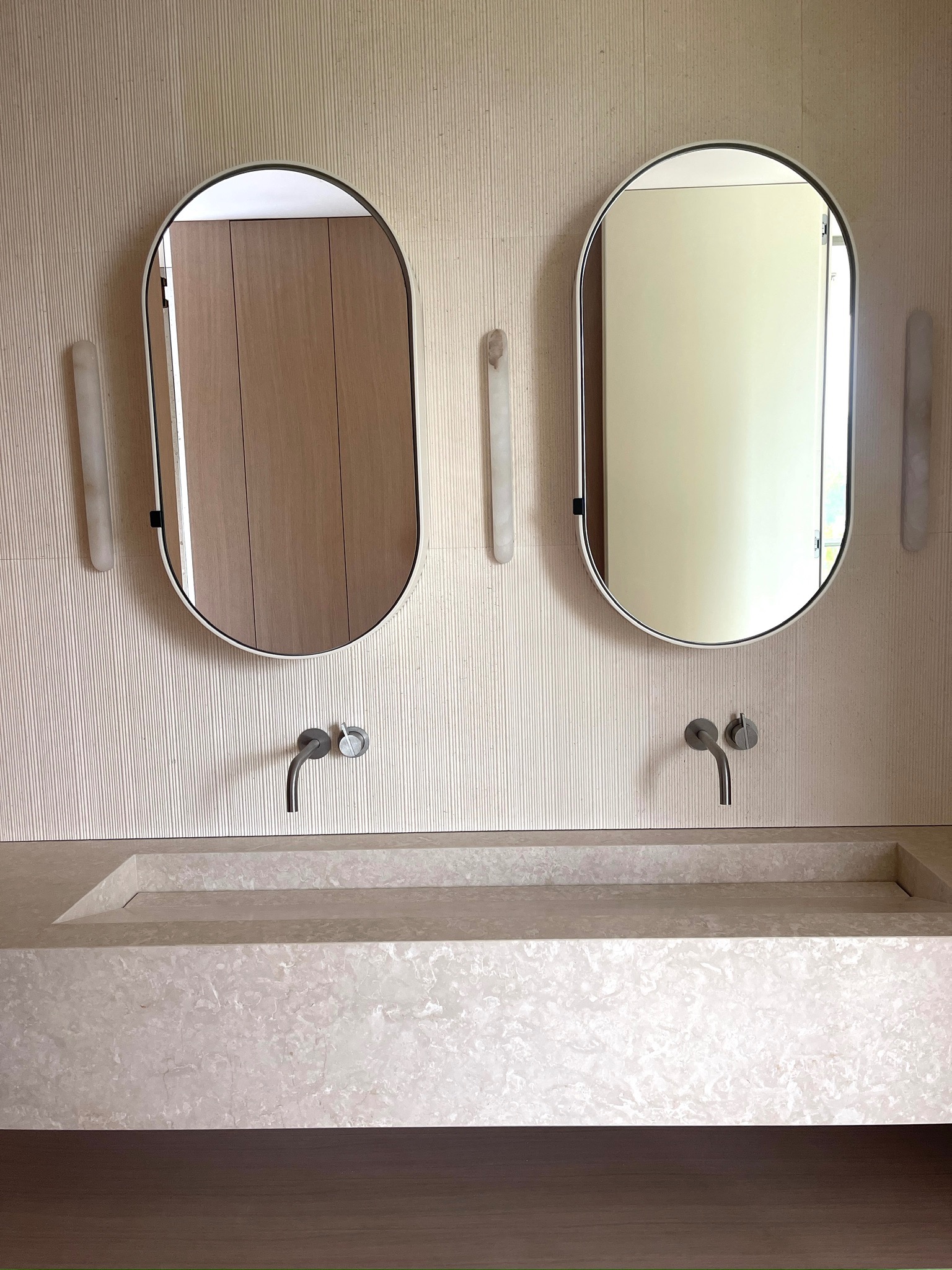 Elegancka łazienka z dwoma owalnymi lustrami, marmurową umywalką i subtelnym oświetleniem ściennym. Minimalistyczny design w jasnych, stonowanych barwach.