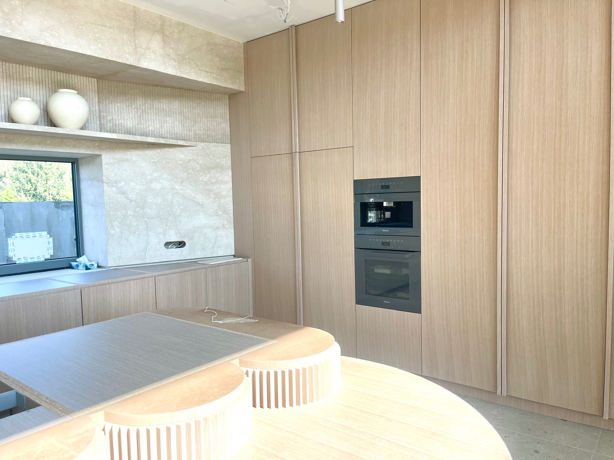 Jasna, nowoczesna kuchnia z drewnianymi szafkami i wyspą w Warszawie. Widoczne wbudowane sprzęty AGD i dekoracyjne wazony na półce.