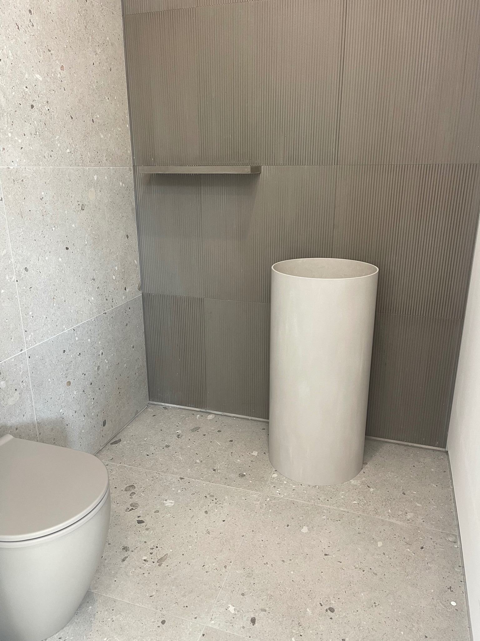 Nowoczesna łazienka z szarymi płytkami lastryko na podłodze i ścianach, oraz pionowo żłobionymi panelami. Widoczna toaleta i cylindryczna umywalka.