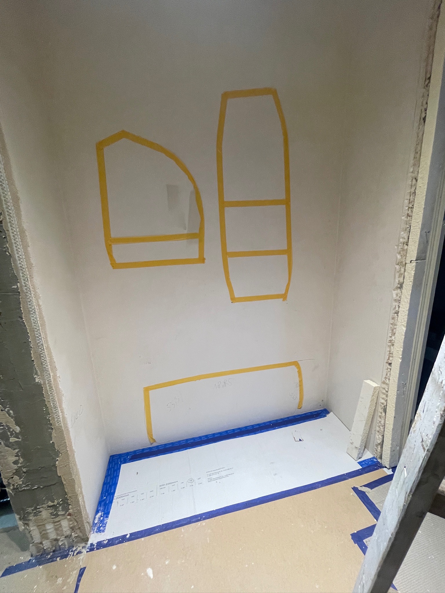 Przygotowanie wnęki: ściany zarysowane taśmą malarską, zabezpieczona podłoga. Etap prac przed wykończeniem, widoczne surowe ściany i drabina.