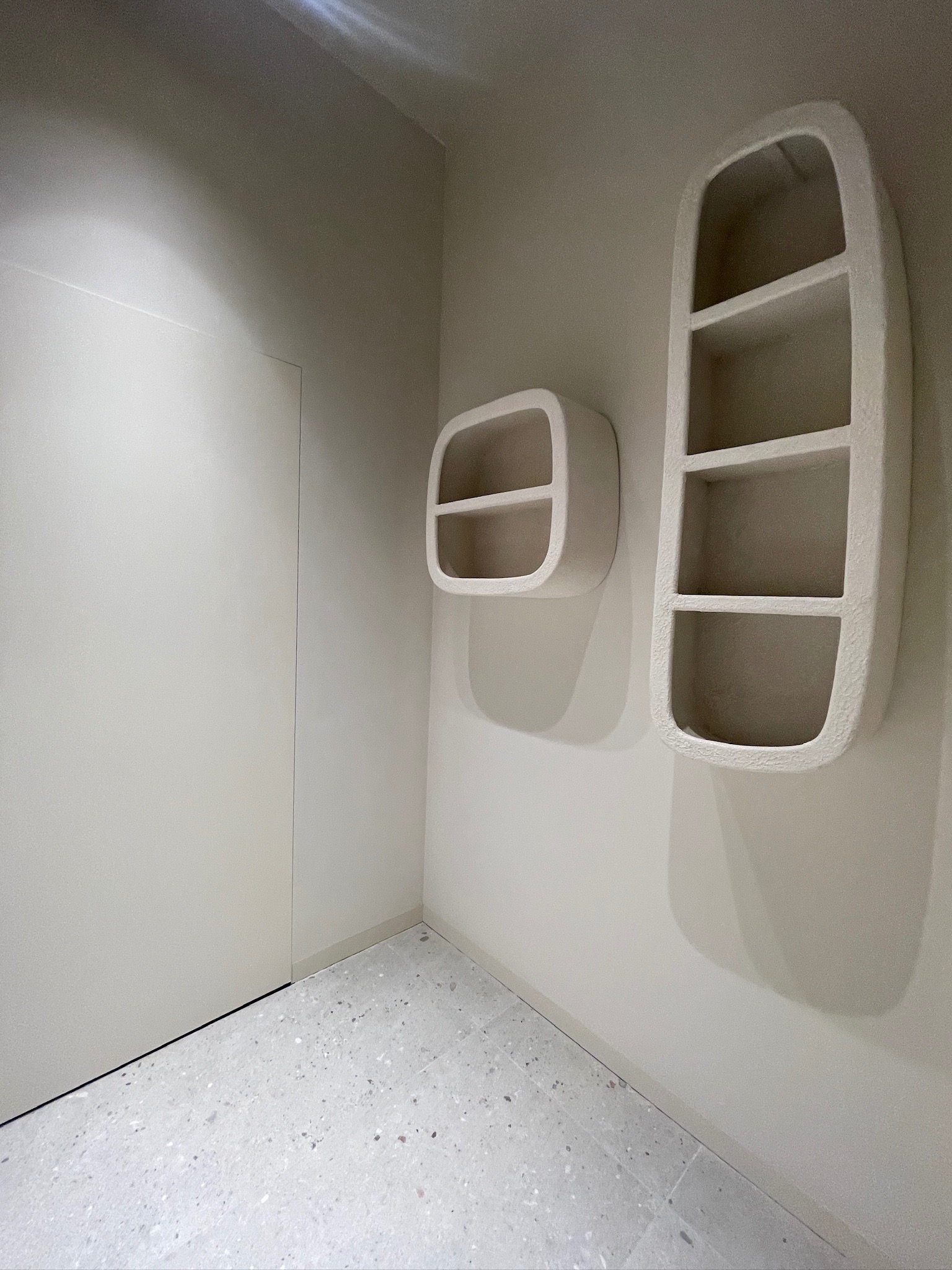 Minimalistyczne wnętrze: kremowe ściany, szara podłoga lastryko i dwie oryginalne, zaokrąglone półki ścienne w stylu artystycznym. Drzwi w kolorze ściany.