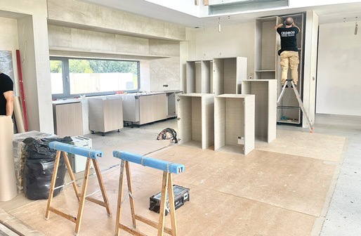 Montaż szafek kuchennych w surowym wnętrzu. Widoczne elementy zabudowy, drabina i narzędzia. Pracownik na drabinie montuje górne szafki. Jasne, naturalne światło.