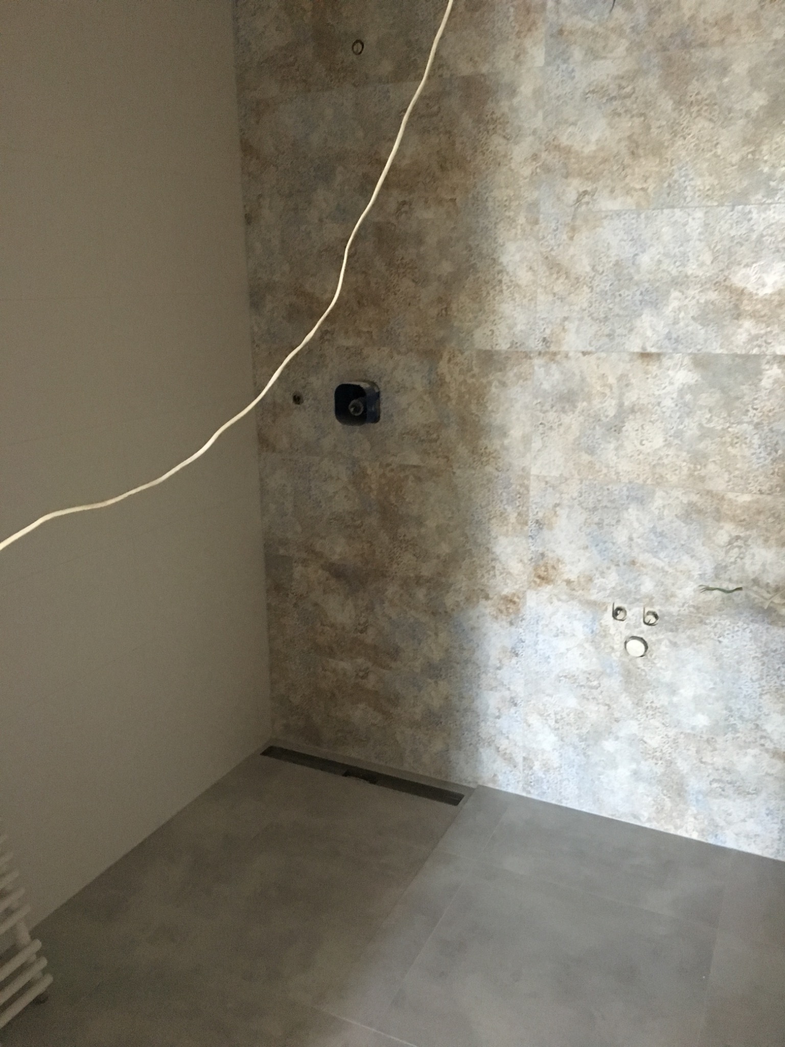 Wykończona łazienka z beżowo-szarą mozaiką na ścianie, widoczne przyłącza wodne i odpływ liniowy w podłodze. Kabel elektryczny zwisa ze ściany.