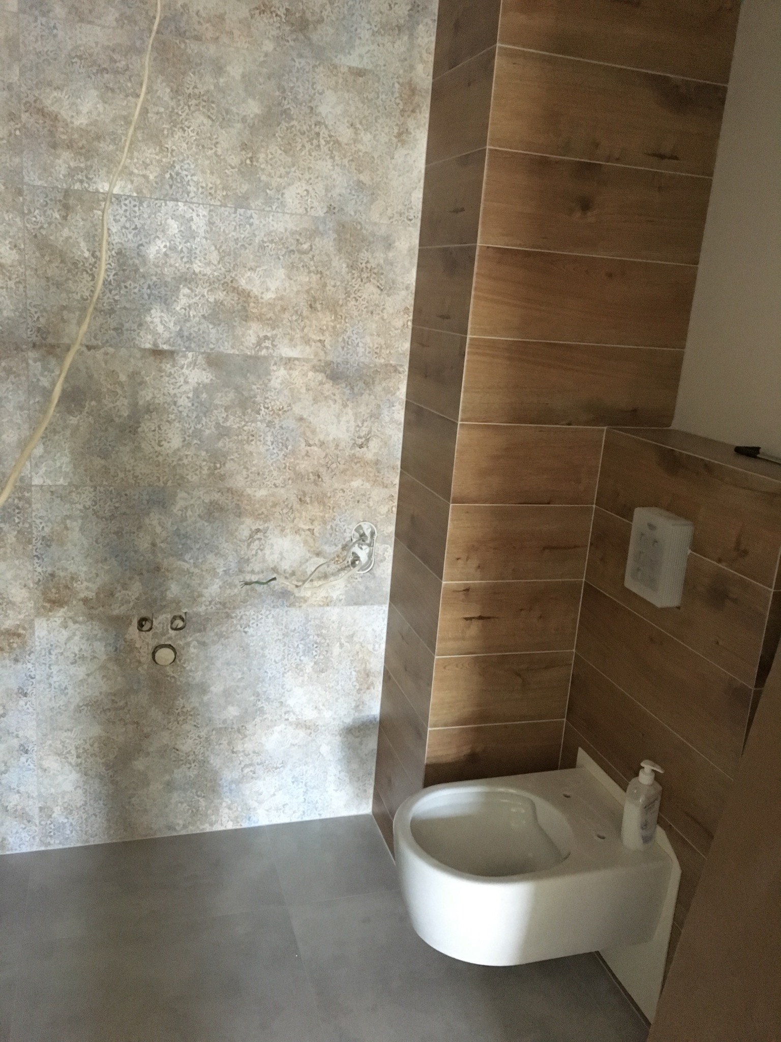 Wykończona łazienka z drewnopodobnymi płytkami na ścianie i podwieszaną toaletą. Widoczne przyłącza elektryczne i hydrauliczne. Szare płytki podłogowe.