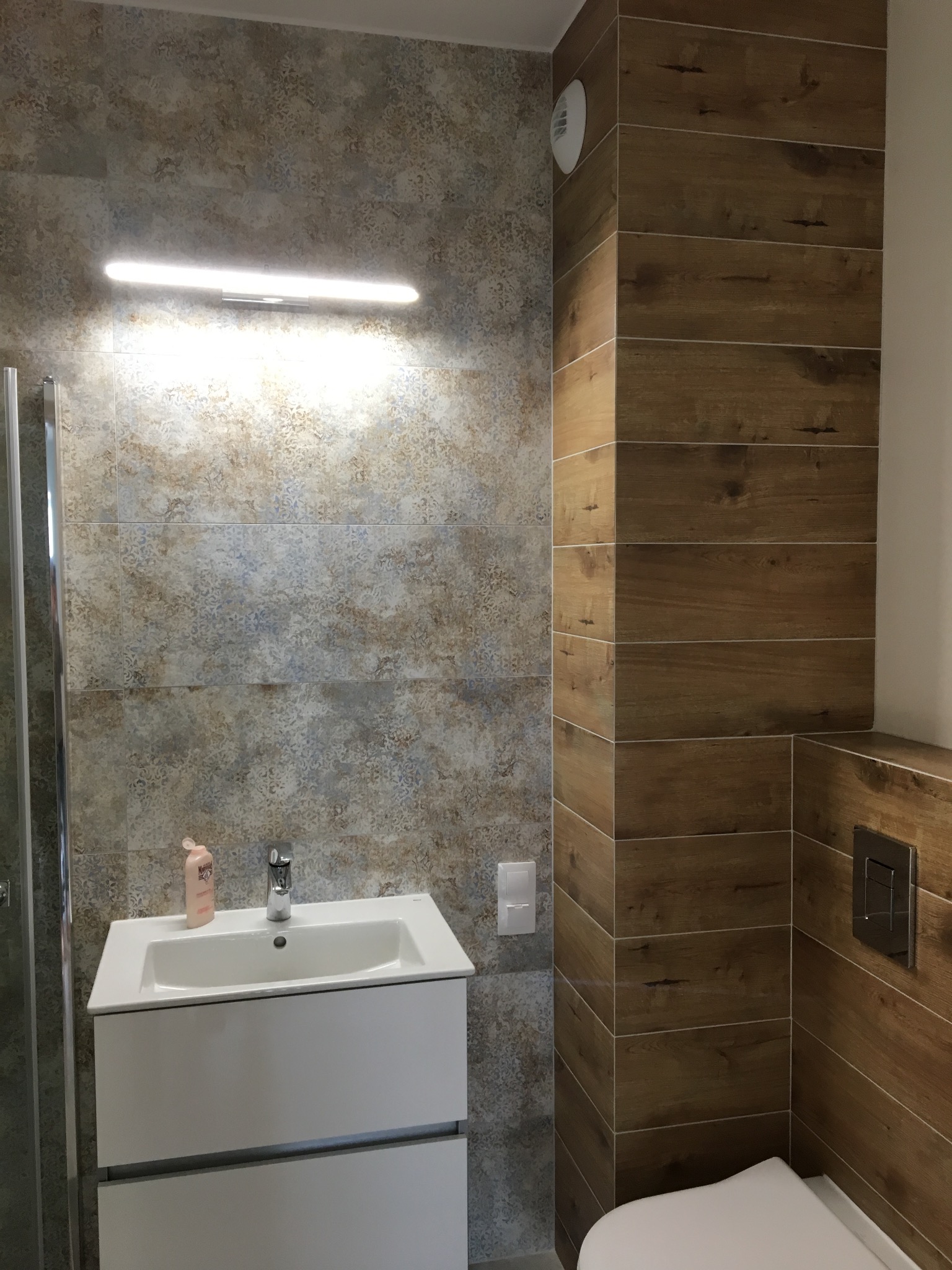 Nowoczesna łazienka po remoncie: biała szafka z umywalką, podświetlane lustro, płytki imitujące drewno i kamień, minimalistyczna armatura, detale wykończeniowe.