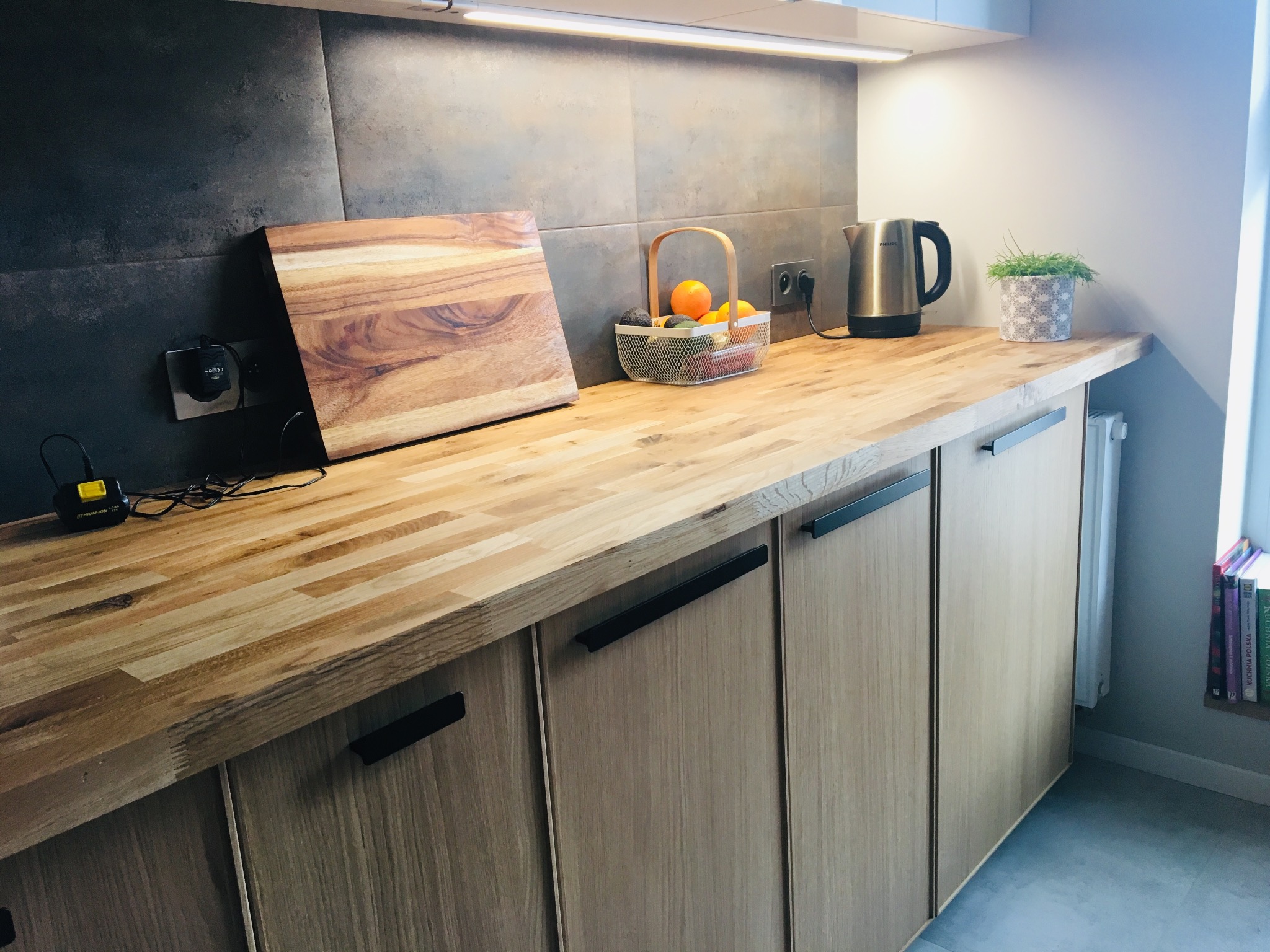 Jasna, drewniana zabudowa kuchenna z czarnymi uchwytami, szarymi płytkami na ścianie i drewnianą deską do krojenia. Minimalistyczny styl, nowoczesne wykończenie.