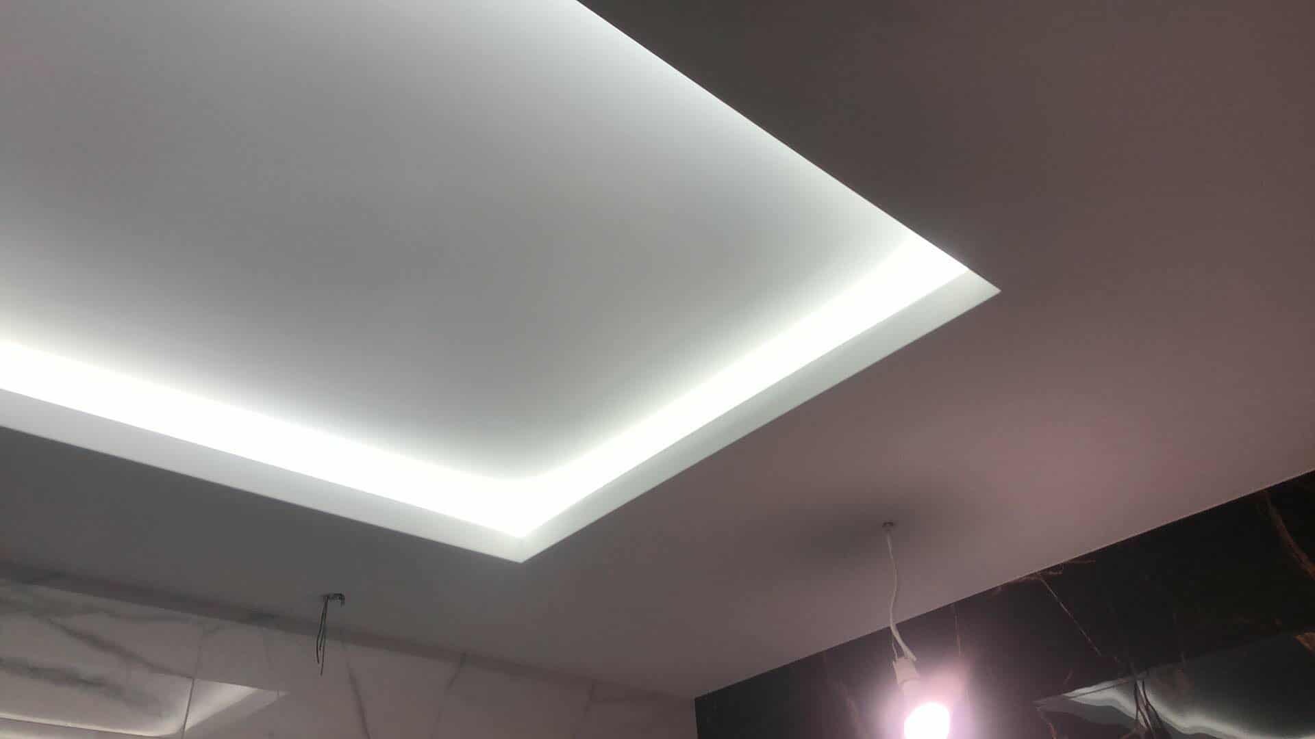 Nowoczesny sufit podwieszany z oświetleniem LED w kształcie prostokąta, widoczne przewody i żarówka, marmurowe wykończenie ściany w tle.