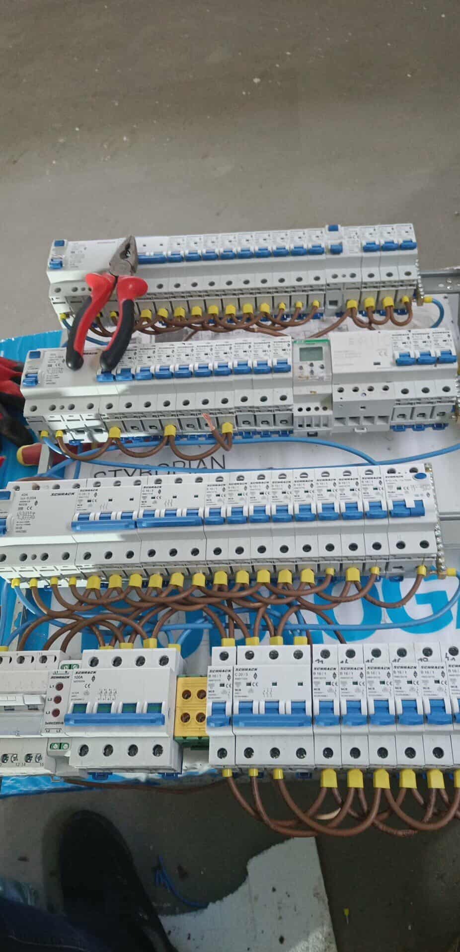 Rozdzielnica elektryczna z bezpiecznikami, brązowymi przewodami i czerwonymi kombinerkami. Widok z góry na instalację elektryczną w trakcie montażu.