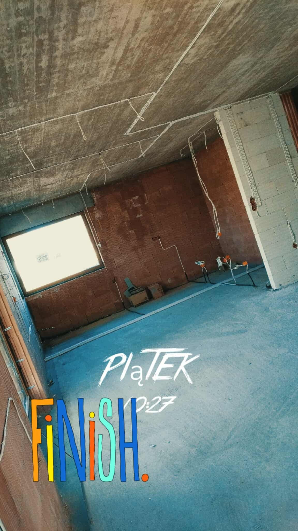 Surowy stan wnętrza z widoczną instalacją elektryczną na suficie i ścianach, przed tynkowaniem. Okno w tle, ceglane ściany. Widoczne kable i puszki.