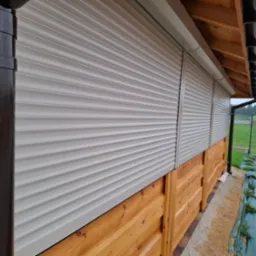 Zamknięte rolety zewnętrzne w kolorze jasnoszarym na drewnianej elewacji budynku, widoczne rynny. Ujęcie z dołu, podkreślające detale konstrukcyjne.