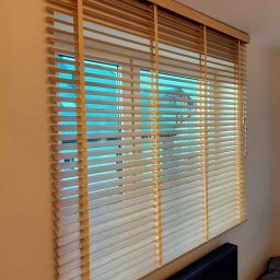 SAW-ROL rolety bramy moskitiery - Drewniane żaluzje poziome w oknie, z widokiem na błękitne dachy. Grzejnik pod oknem. Naturalne światło podkreśla strukturę drewna.