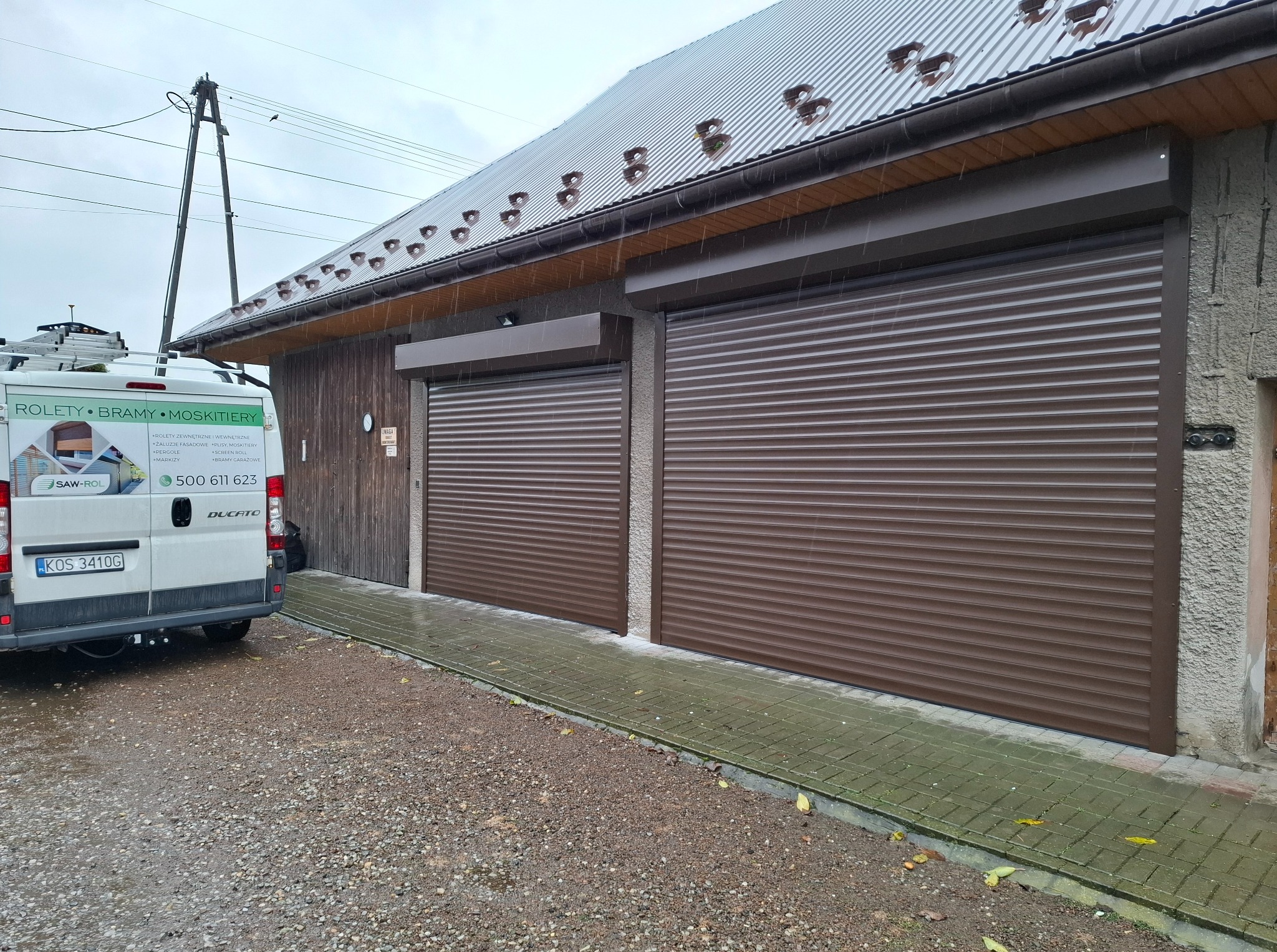 Dwie brązowe rolety garażowe zewnętrzne, zamontowane w budynku z szarą elewacją. Obok zaparkowany biały bus z reklamą rolet i bram. Deszczowa pogoda.