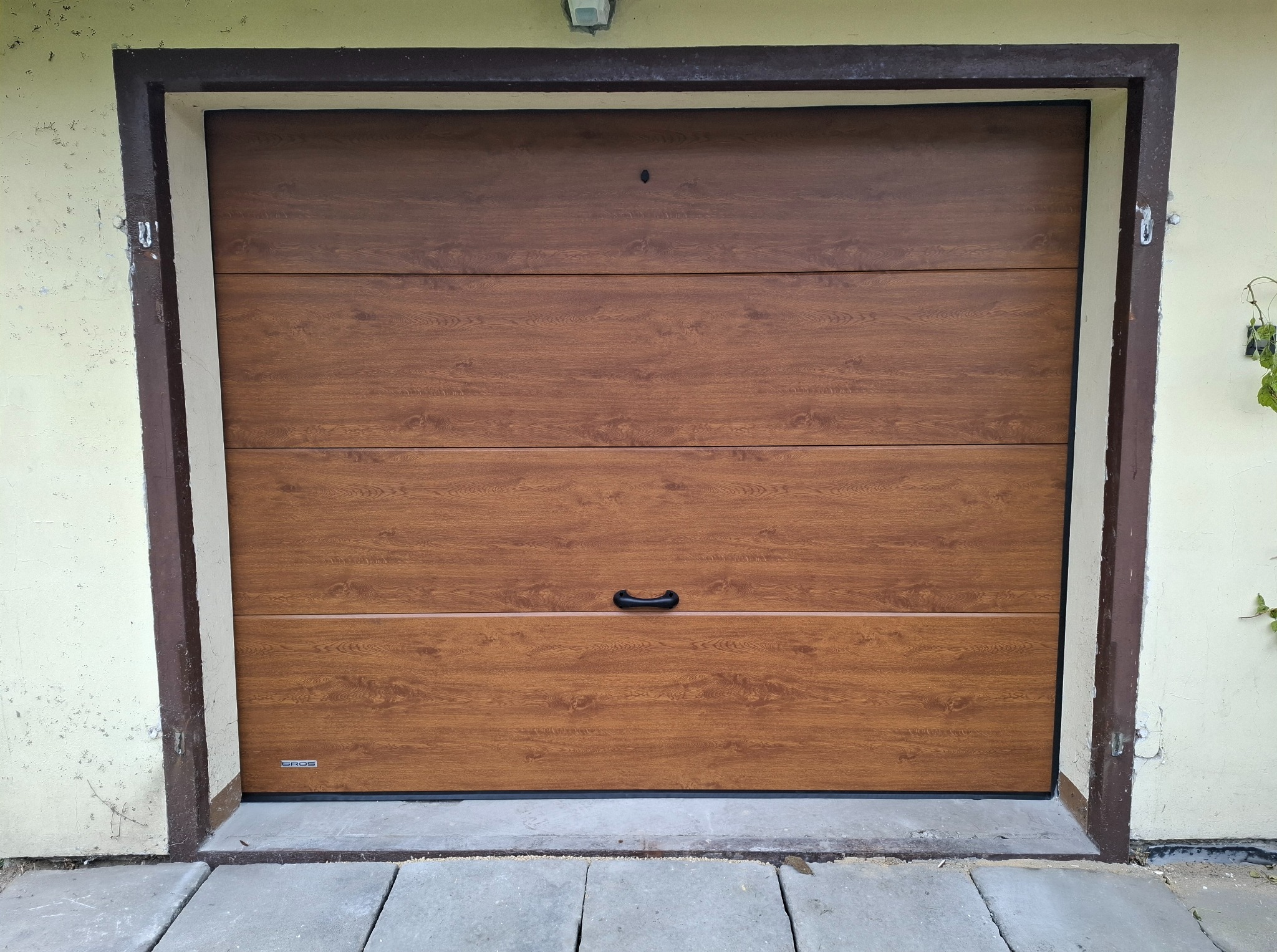 Zamknięta, segmentowa brama garażowa w odcieniach brązu, imitująca drewno, z czarnym uchwytem, zamontowana w jasnym budynku. Widoczne detale wykończenia.