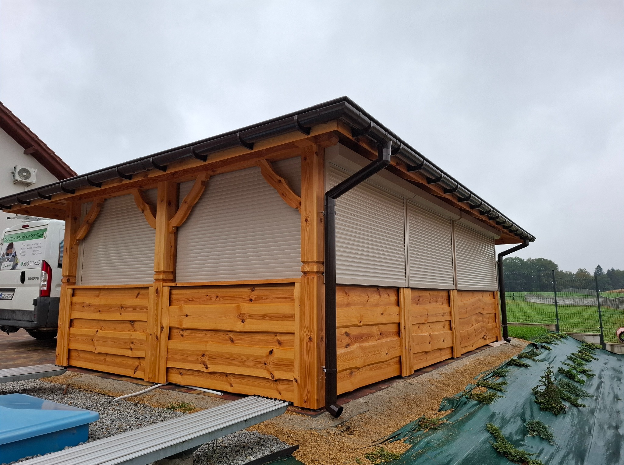 Drewniana altana z roletami zewnętrznymi w kolorze beżowym, z brązowymi rynnami. Widoczny fragment dachu domu i samochód dostawczy w tle. Pochmurny dzień.