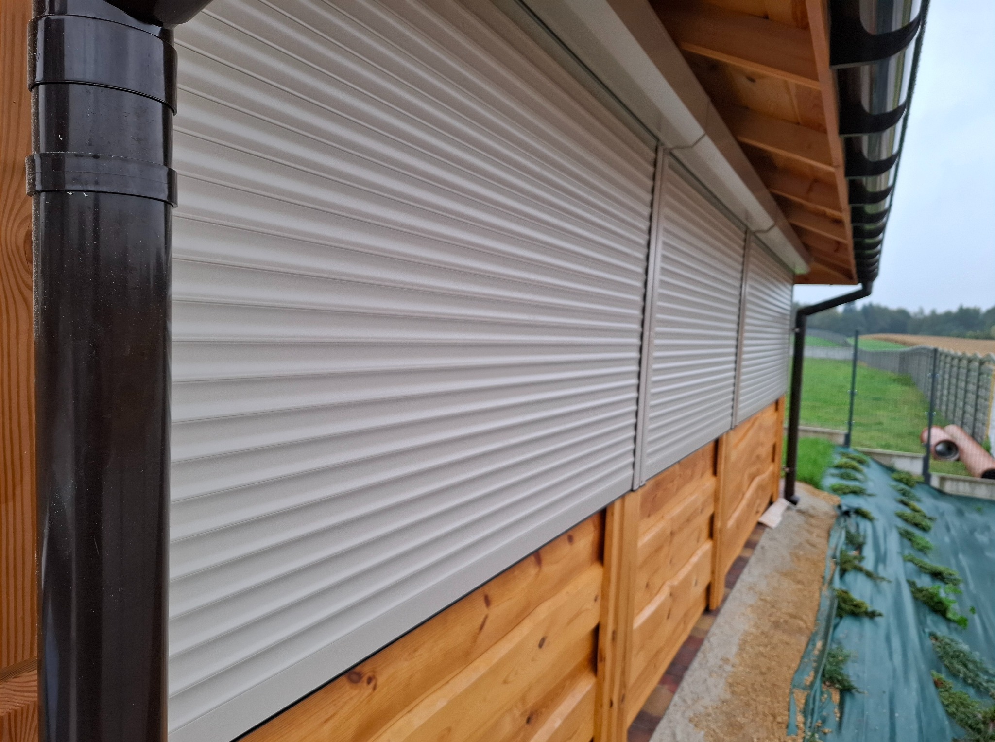 Zamknięte rolety zewnętrzne w kolorze jasnoszarym na drewnianej elewacji budynku, widoczne rynny. Ujęcie z dołu, podkreślające detale konstrukcyjne.