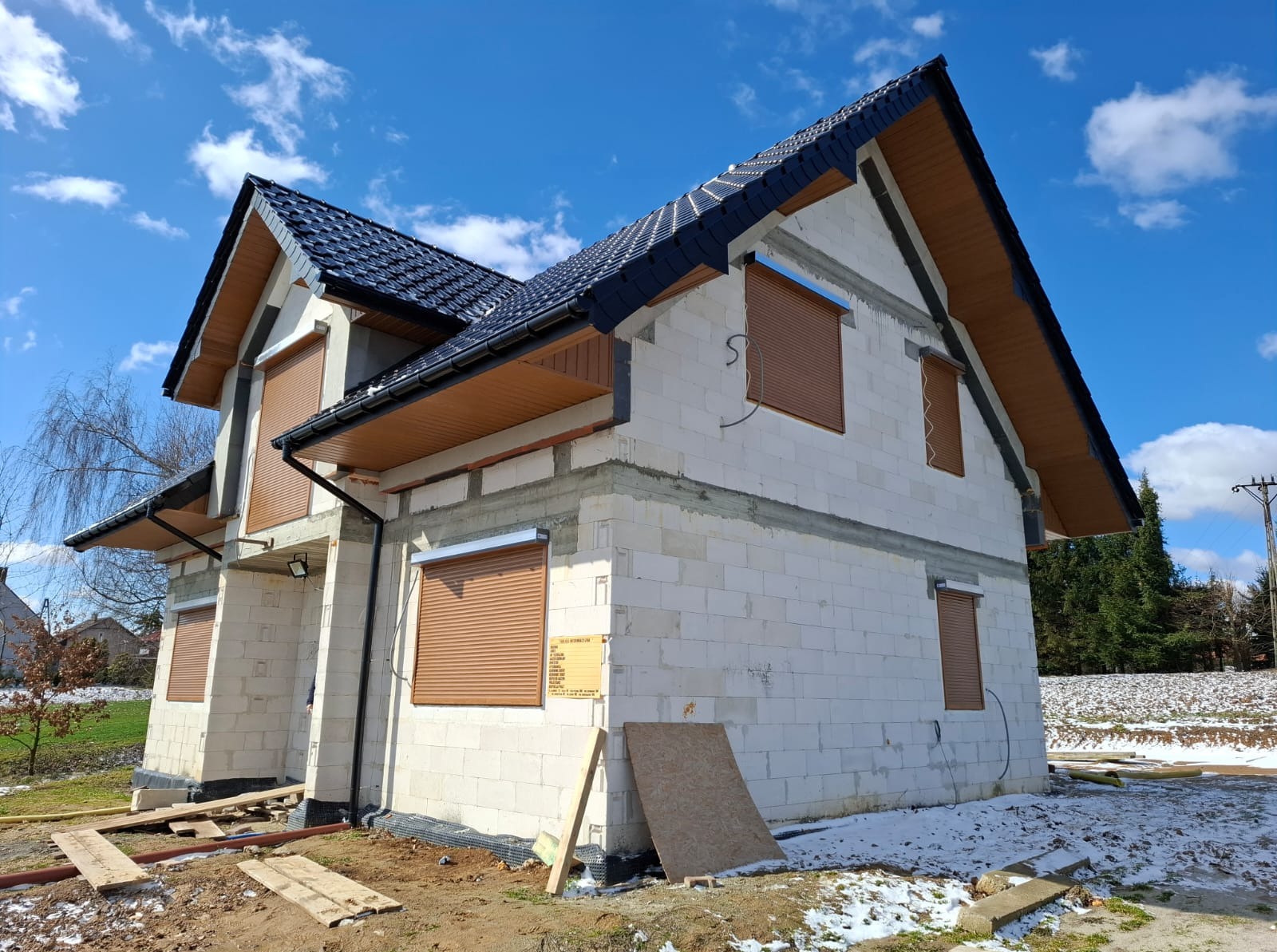 Dom w trakcie budowy z zamontowanymi brązowymi roletami zewnętrznymi. Widoczne bloczki betonowe, czarny dach i błękitne niebo z chmurami w tle.