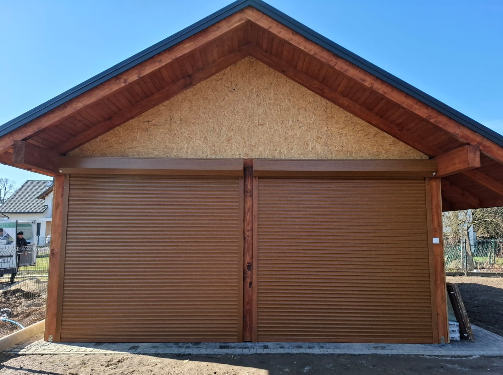 Zewnętrzne, brązowe rolety garażowe w drewnianej konstrukcji z widocznym zarysem budynku w tle. Ujęcie z poziomu gruntu, w słoneczny dzień.