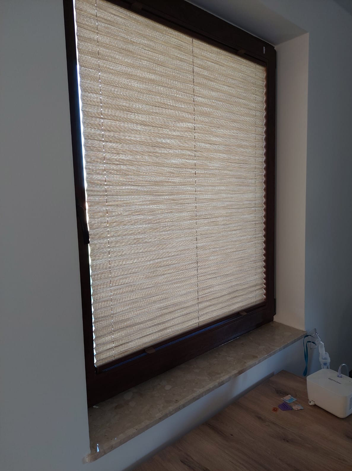 Beżowa roleta wewnętrzna plisowana w brązowej ramie okiennej, zamontowana w pokoju o szarych ścianach. Na parapecie widoczny sprzęt medyczny.