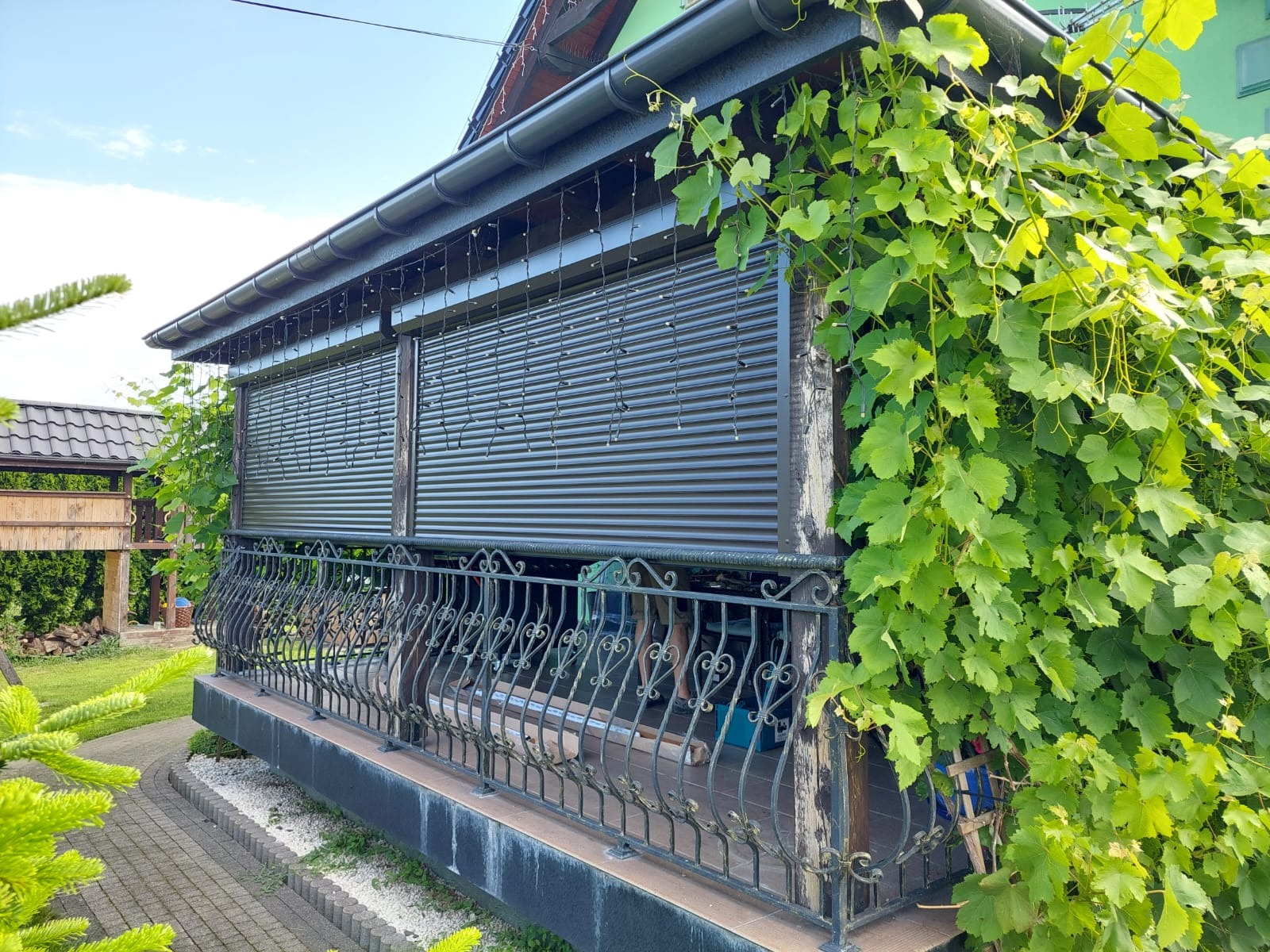 Zadaszony taras z brązowymi roletami zewnętrznymi, ozdobiony pnącą roślinnością i dekoracyjnymi lampkami. Metalowa balustrada z ornamentami.
