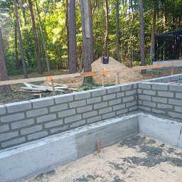ESSENTIS Konrad Joński - Wzniesione ściany fundamentowe z bloczków betonowych na tle leśnym. Widoczne drewniane stemple i piasek. Ujęcie z poziomu gruntu, w słoneczny dzień.