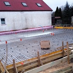 Stanisław Kaczyński - Przygotowanie zbrojenia pod wylewkę fundamentów domu jednorodzinnego. Widoczne szalunki, siatka zbrojeniowa, izolacja termiczna i budynek w tle.