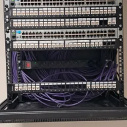 Szafa rack z patch panelem i okablowaniem w kolorze fioletowym. Widoczne porty RJ45 i listwa zasilająca. Górna część szafy wyposażona w wentylatory.