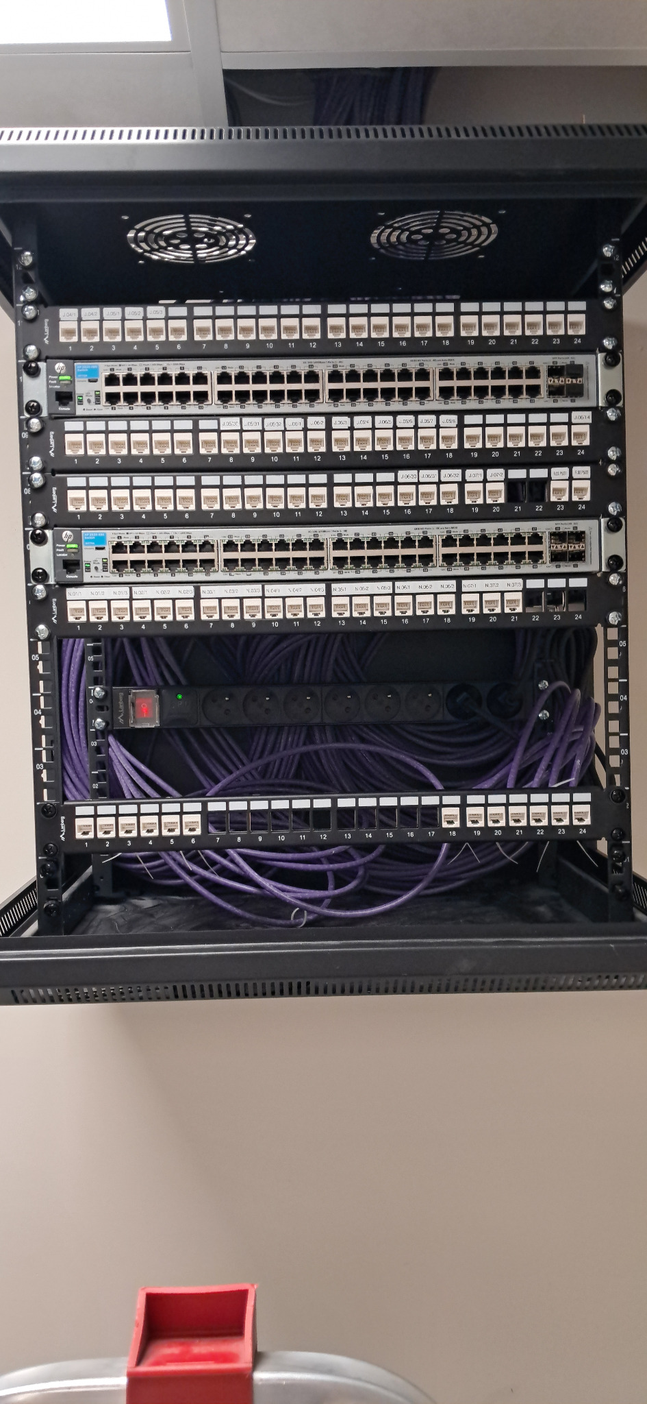 Szafa rack z patch panelem i okablowaniem w kolorze fioletowym. Widoczne porty RJ45 i listwa zasilająca. Górna część szafy wyposażona w wentylatory.