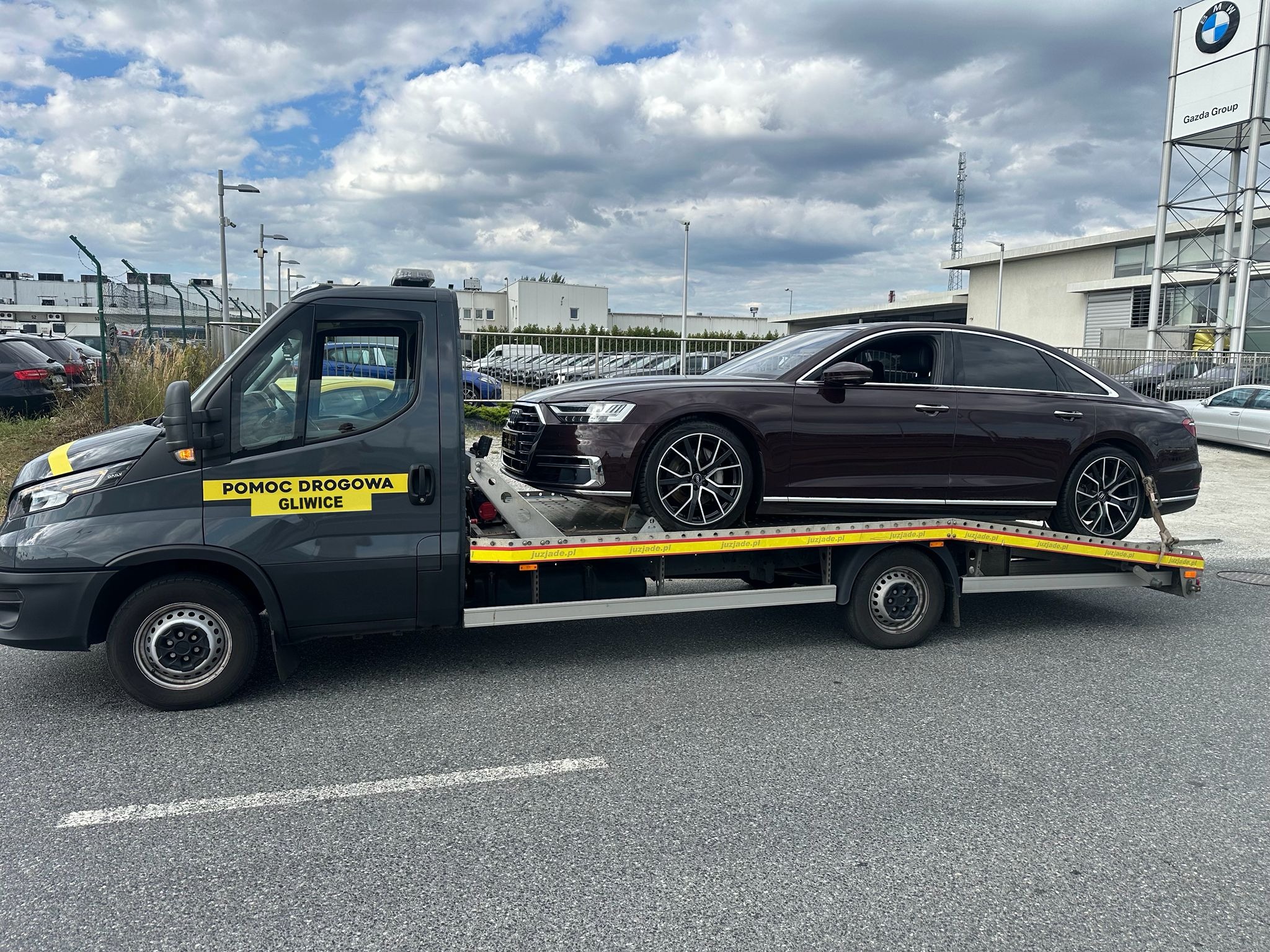 Czarna laweta z napisem 'Pomoc Drogowa Gliwice' transportuje bordowe Audi A8 na tle salonu samochodowego Gazda Group w pochmurny dzień.
