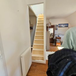 PIN - Jasne, drewniane schody prowadzące na poddasze w jasnym wnętrzu. Widoczny fragment grzejnika i podłogi. Ujęcie z dołu, podkreślające wysokość schodów.