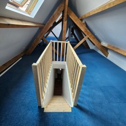 PIN - Adaptacja poddasza: widok z góry na drewniane schody prowadzące do piwnicy, niebieska wykładzina, drewniane belki stropowe, okno dachowe.