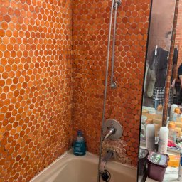 PIN - Pomarańczowa mozaika w kabinie prysznicowej z widocznymi śladami użytkowania i uszkodzeniami fugi przy baterii. W tle lustro z odbiciem osoby.