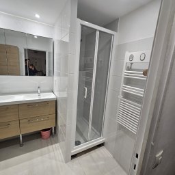 PIN - Jasna łazienka po remoncie: kabina prysznicowa z białymi ramami, szafka z umywalką z drewnianymi frontami, grzejnik drabinkowy. Widoczne białe płytki na ścianach i podłodze.