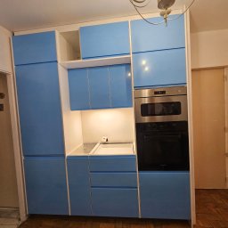 PIN - Niebieska zabudowa kuchenna z piekarnikiem i oświetleniem LED, widok na fragment wnętrza w trakcie montażu, z elementami wykończeniowymi i drewnianą podłogą.