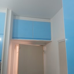 PIN - Niebieskie szafki nad otwartymi drzwiami w białej wnęce, widok z dołu. Minimalistyczny design, jasne kolory, nowoczesne wykończenie wnętrza.