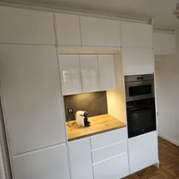 Biała, nowoczesna zabudowa kuchenna z piekarnikiem i ekspresem do kawy na drewnianym blacie, oświetlona punktowo, z widoczną podłogą z cegły.