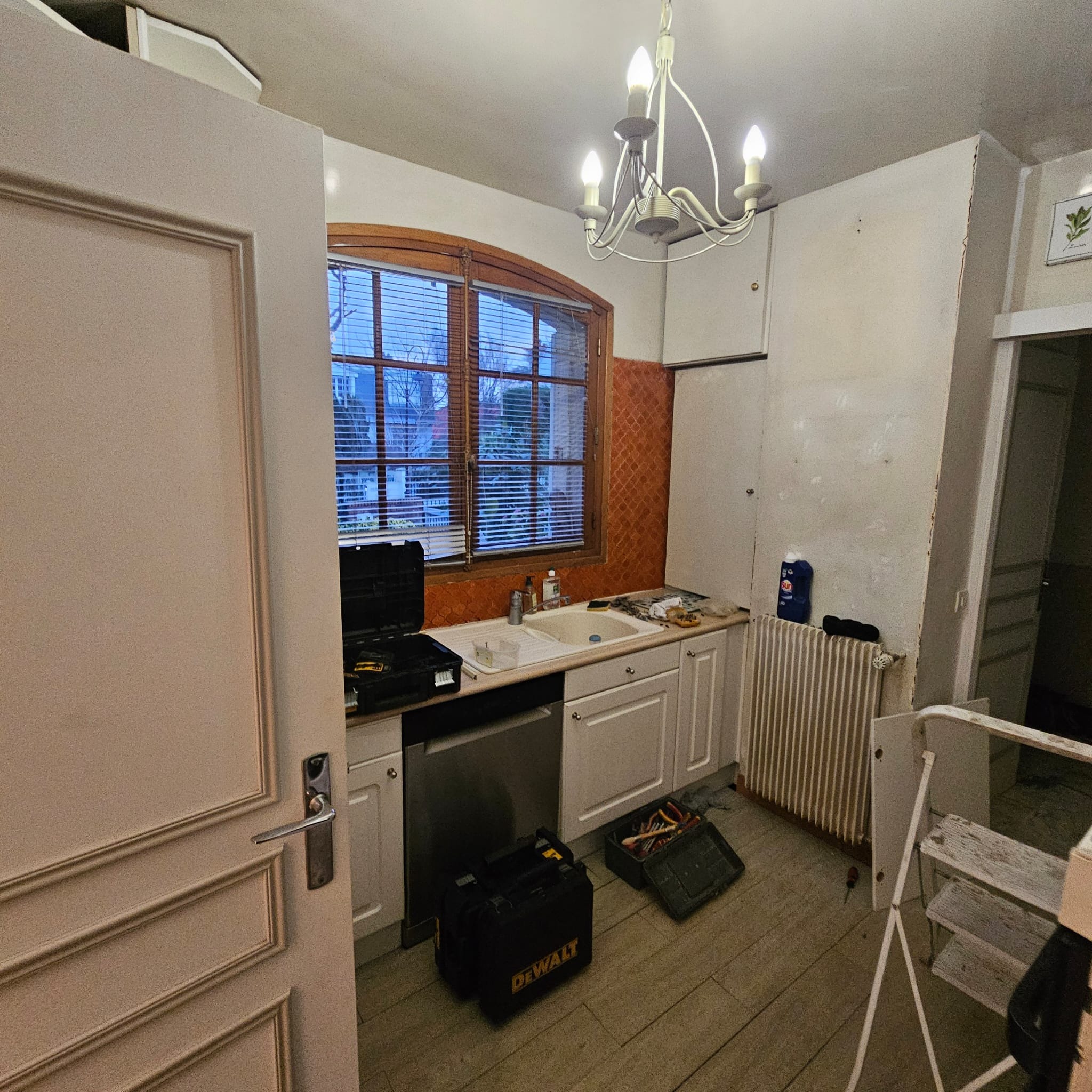 Kuchnia w trakcie remontu: widoczne narzędzia DeWalt, odsłonięte ściany, okno z żaluzjami i wiszący żyrandol. Etap prac wykończeniowych.