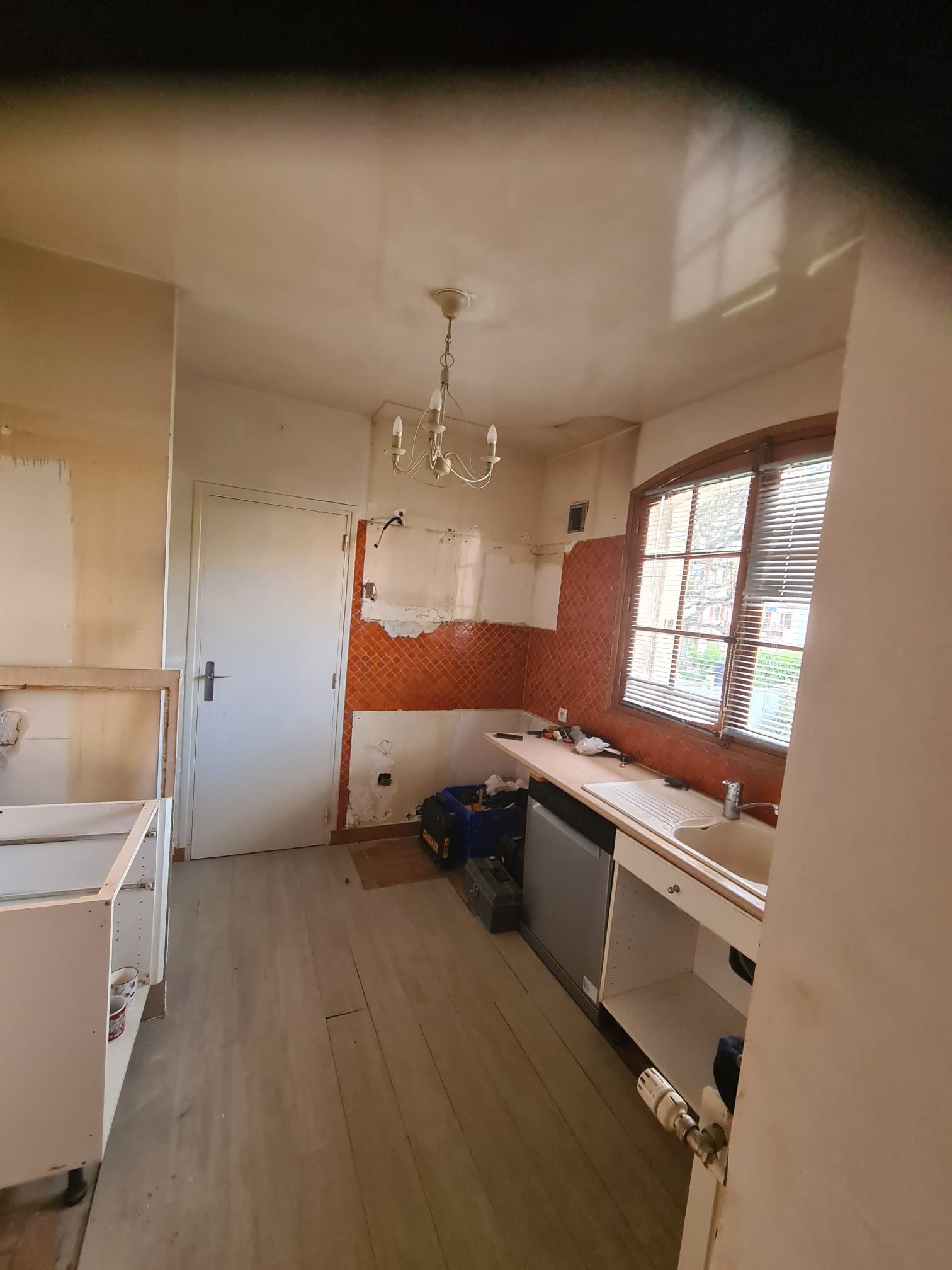 Kuchnia w trakcie remontu: częściowo zdemontowane szafki, odsłonięte fragmenty ściany z pomarańczowymi płytkami, widoczne narzędzia i instalacje. Jasne drewniane panele podłogowe.