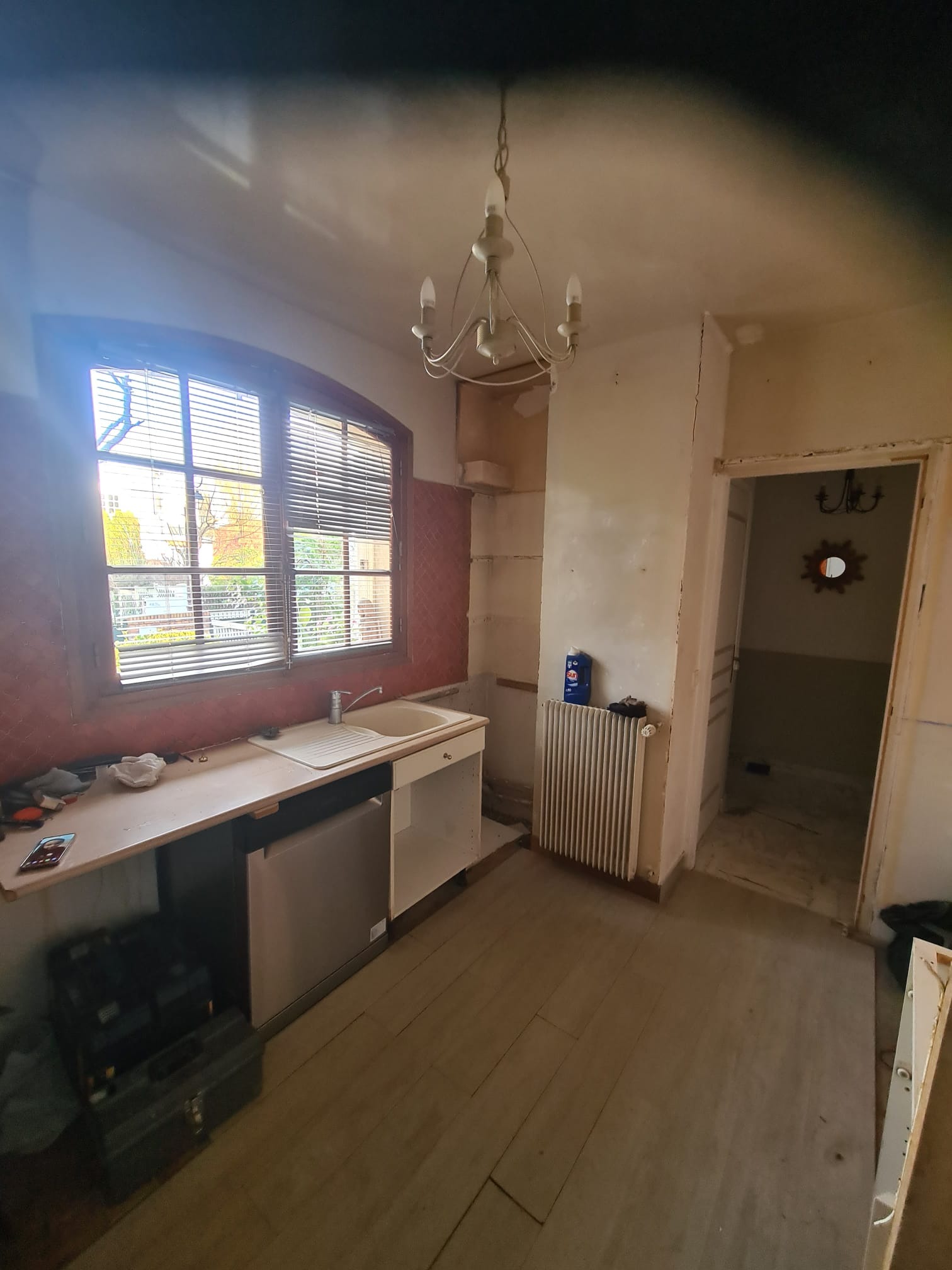 Wnętrze kuchni w trakcie remontu: widoczne stare meble kuchenne, okno z żaluzjami, grzejnik i otwarte drzwi do innego pomieszczenia. Ściany i podłoga wymagają odnowienia.