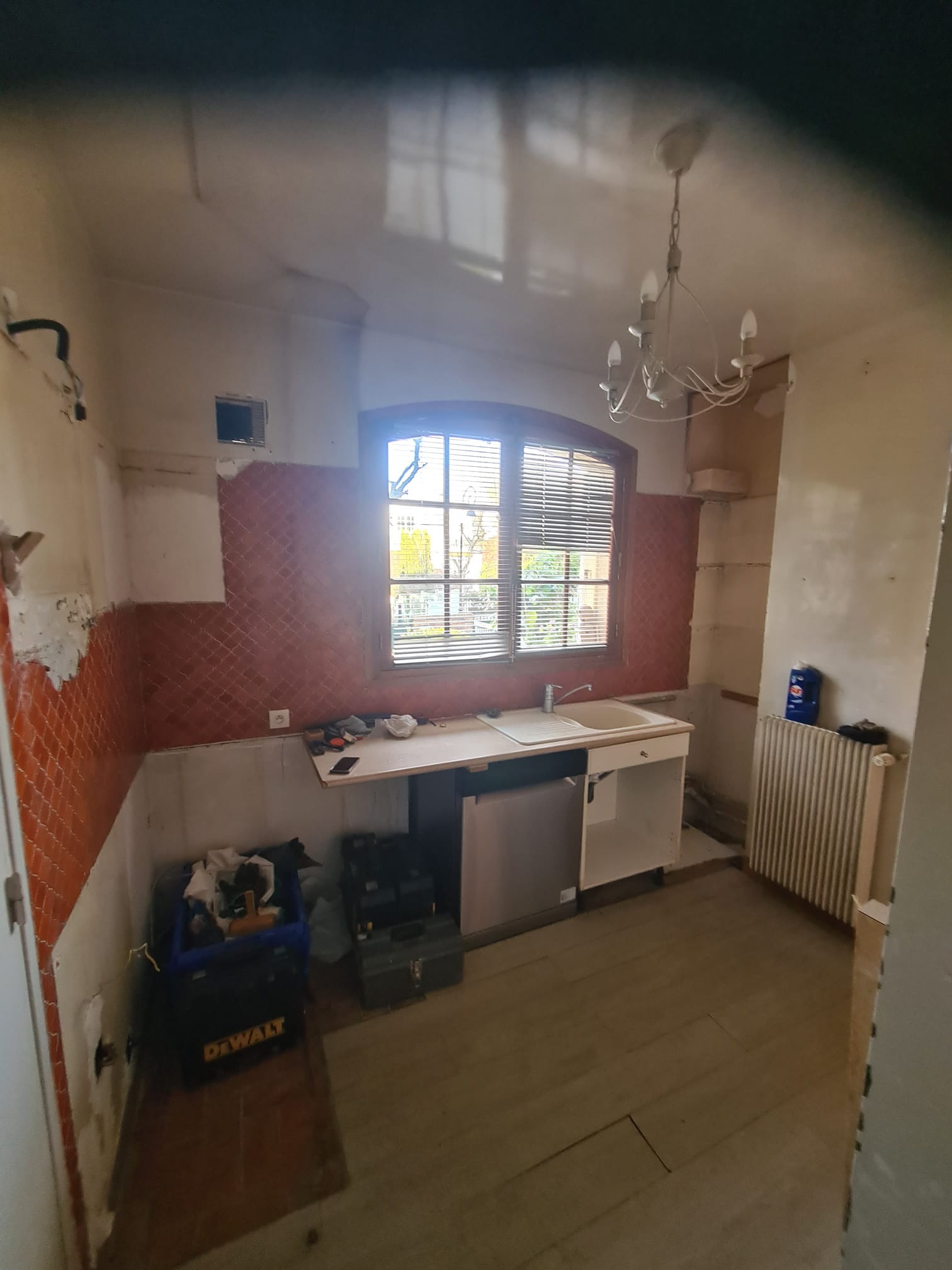 Kuchenne wnętrze w trakcie demontażu płytek i przygotowania do remontu. Widoczne narzędzia, odsłonięte ściany i okno z widokiem na zewnątrz.