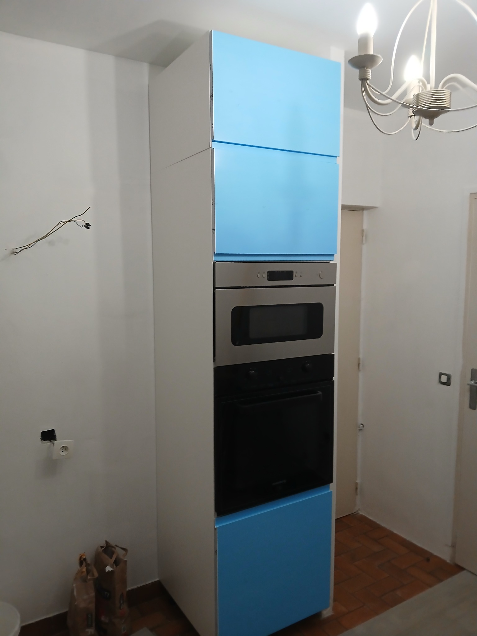 Wysoka zabudowa kuchenna z niebieskimi frontami i sprzętem AGD w Nysie. Widoczne niedokończone prace elektryczne i worki z zaprawą.