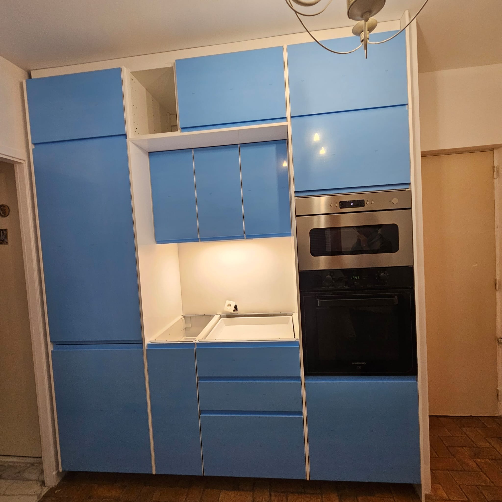 Niebieska zabudowa kuchenna z piekarnikiem i oświetleniem LED, widok na fragment wnętrza w trakcie montażu, z elementami wykończeniowymi i drewnianą podłogą.