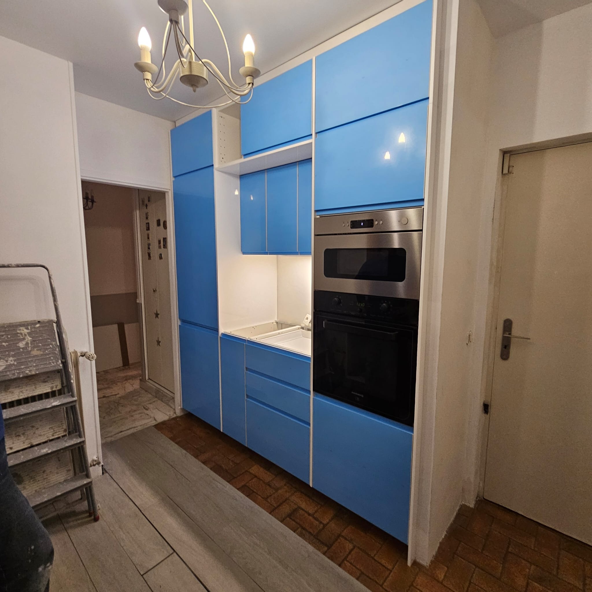 Niebieska zabudowa kuchenna z piekarnikiem i mikrofalówką, widok na fragment podłogi z dwoma rodzajami płytek, drabina malarska w rogu.