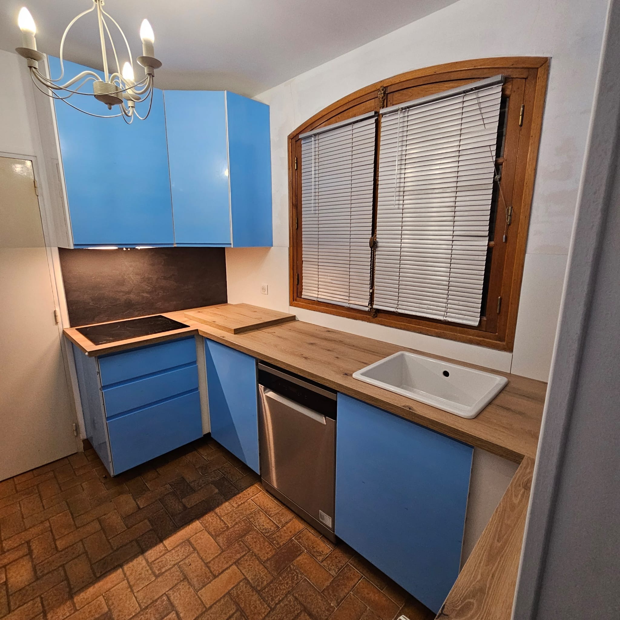 Niebieska zabudowa kuchenna z drewnianym blatem i oknem z żaluzjami. Widoczna płyta indukcyjna, zlew i zmywarka. Podłoga wyłożona ceglastą terakotą.