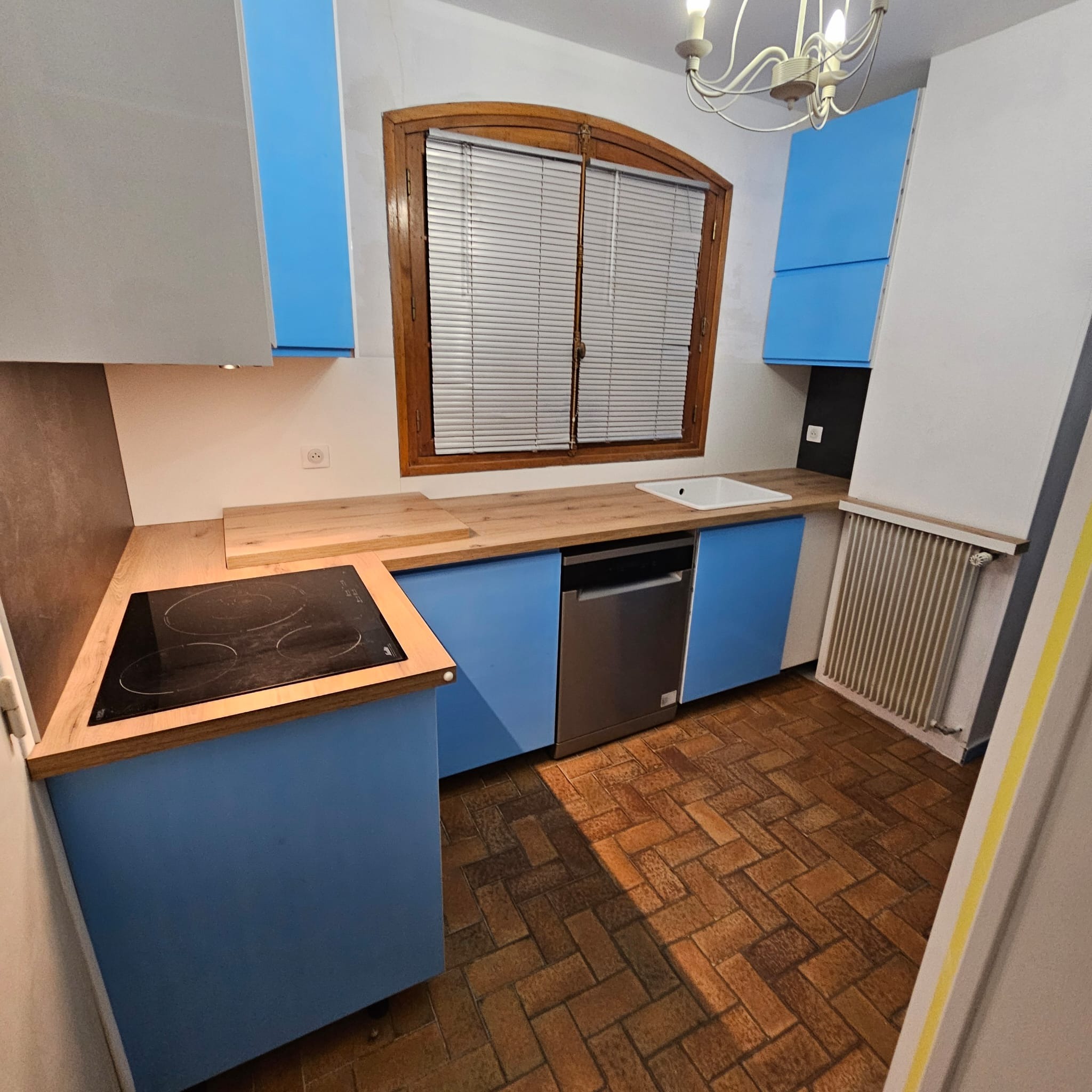 Niebieska zabudowa kuchenna z drewnianym blatem, widok na płytę indukcyjną, zlew i okno z żaluzjami. Brązowa podłoga z cegiełką. Nowoczesny design.