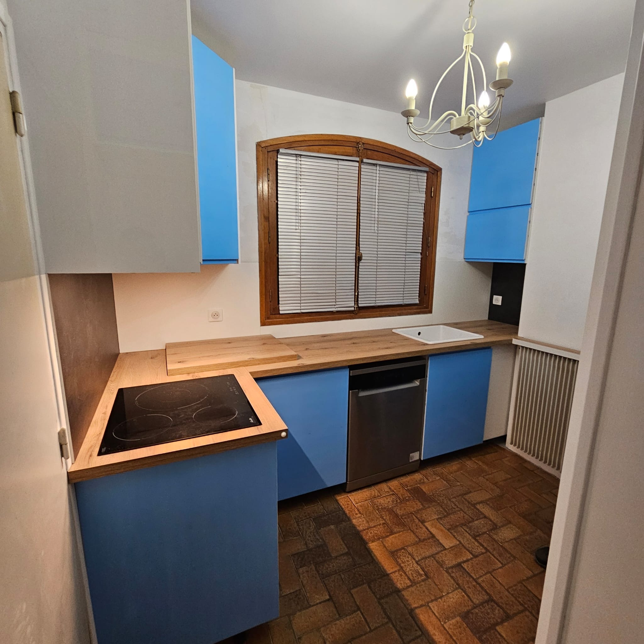 Niebieska zabudowa kuchenna z drewnianym blatem i oknem w brązowej ramie. Widoczna płyta indukcyjna, zlew i parkiet ułożony w jodełkę. Całość w jasnym pomieszczeniu.