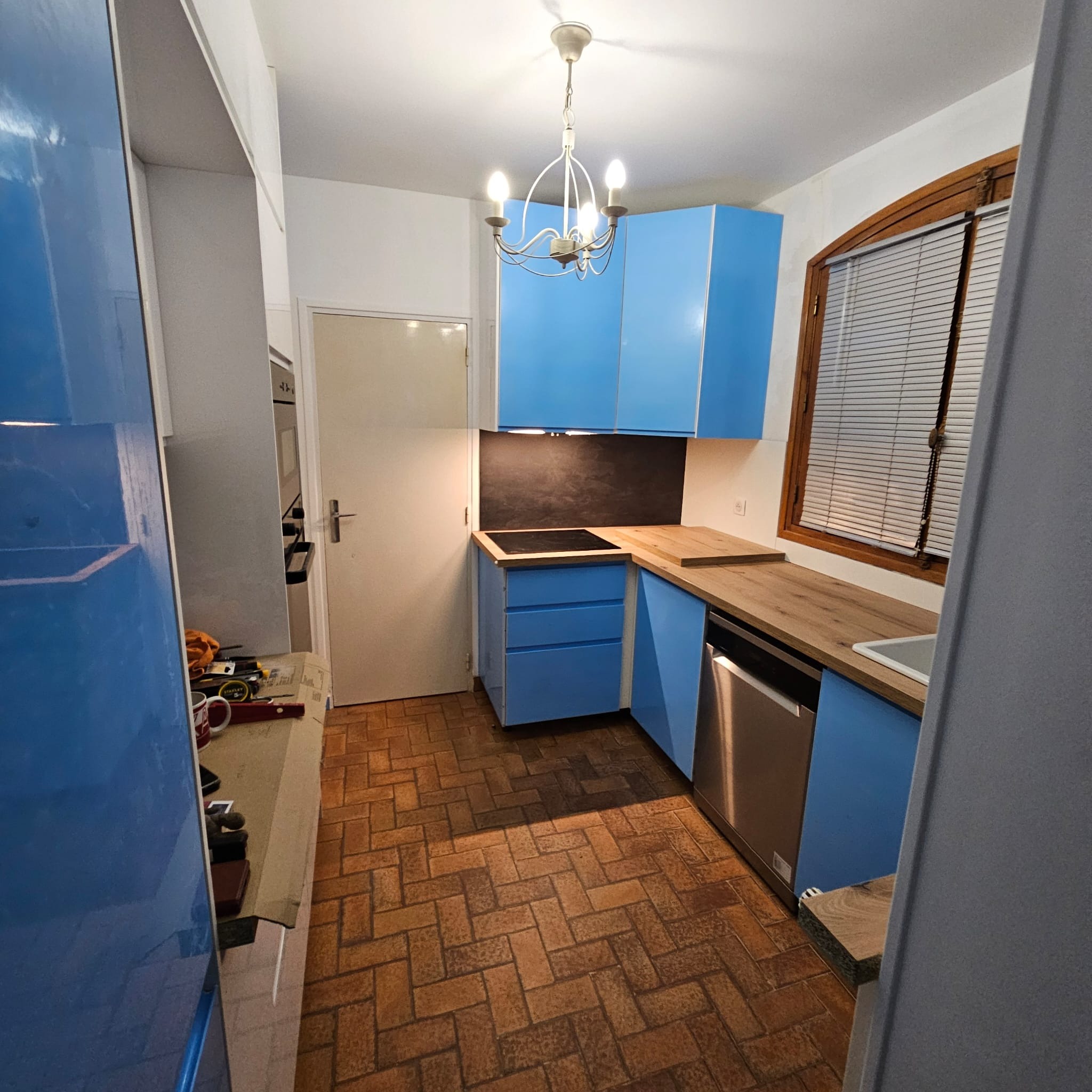 Niebieska zabudowa kuchenna z drewnianym blatem i oknem z żaluzjami. Podłoga z brązowej cegły. Widoczny fragment stołu z narzędziami.